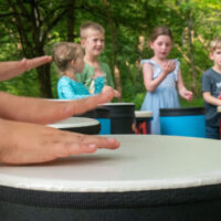 Young kid plays a drum with others in the background passing a shaker egg in a drum circle.